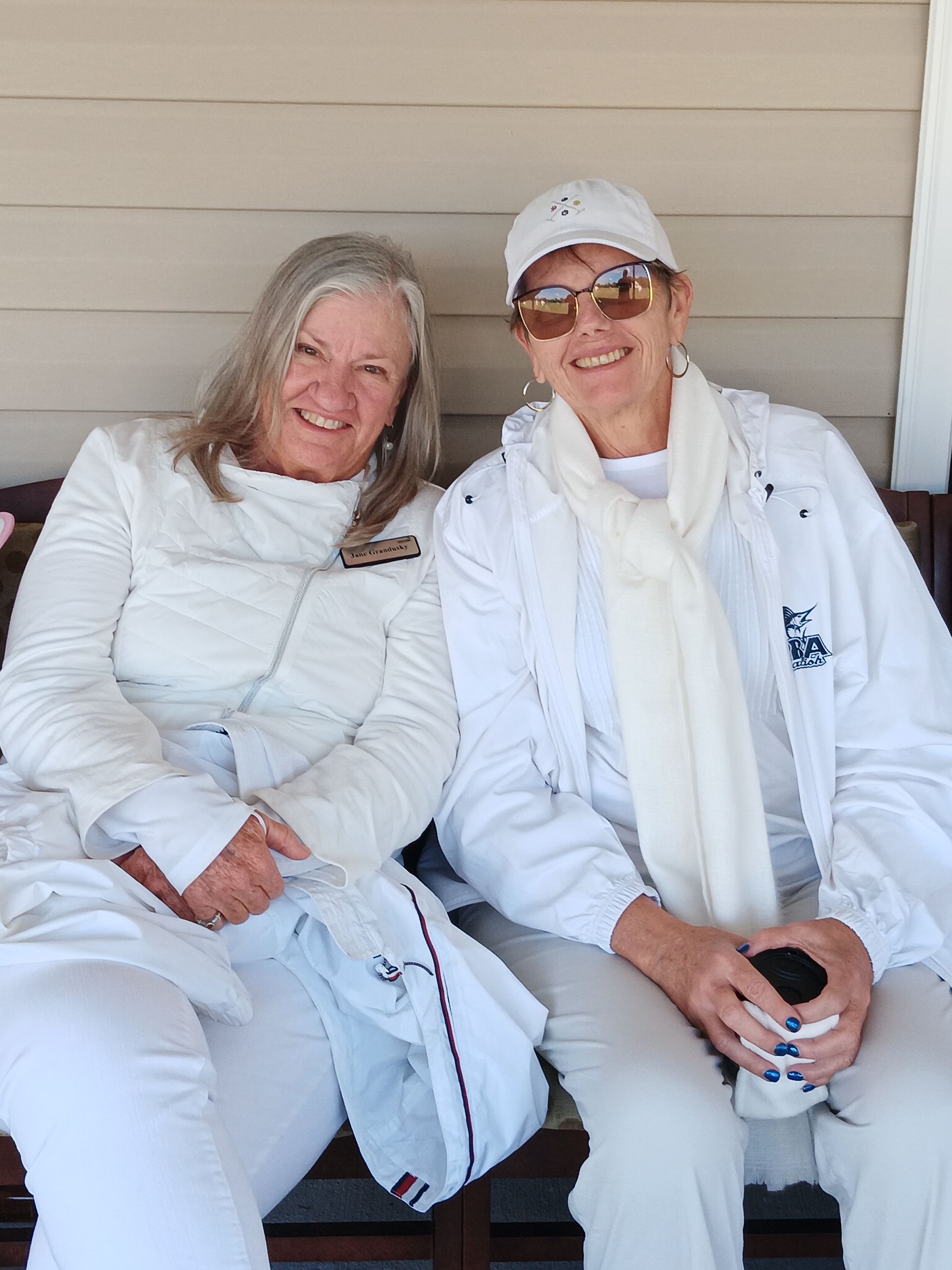 Two women in white outfits smiling, seated on a bench, outdoors.