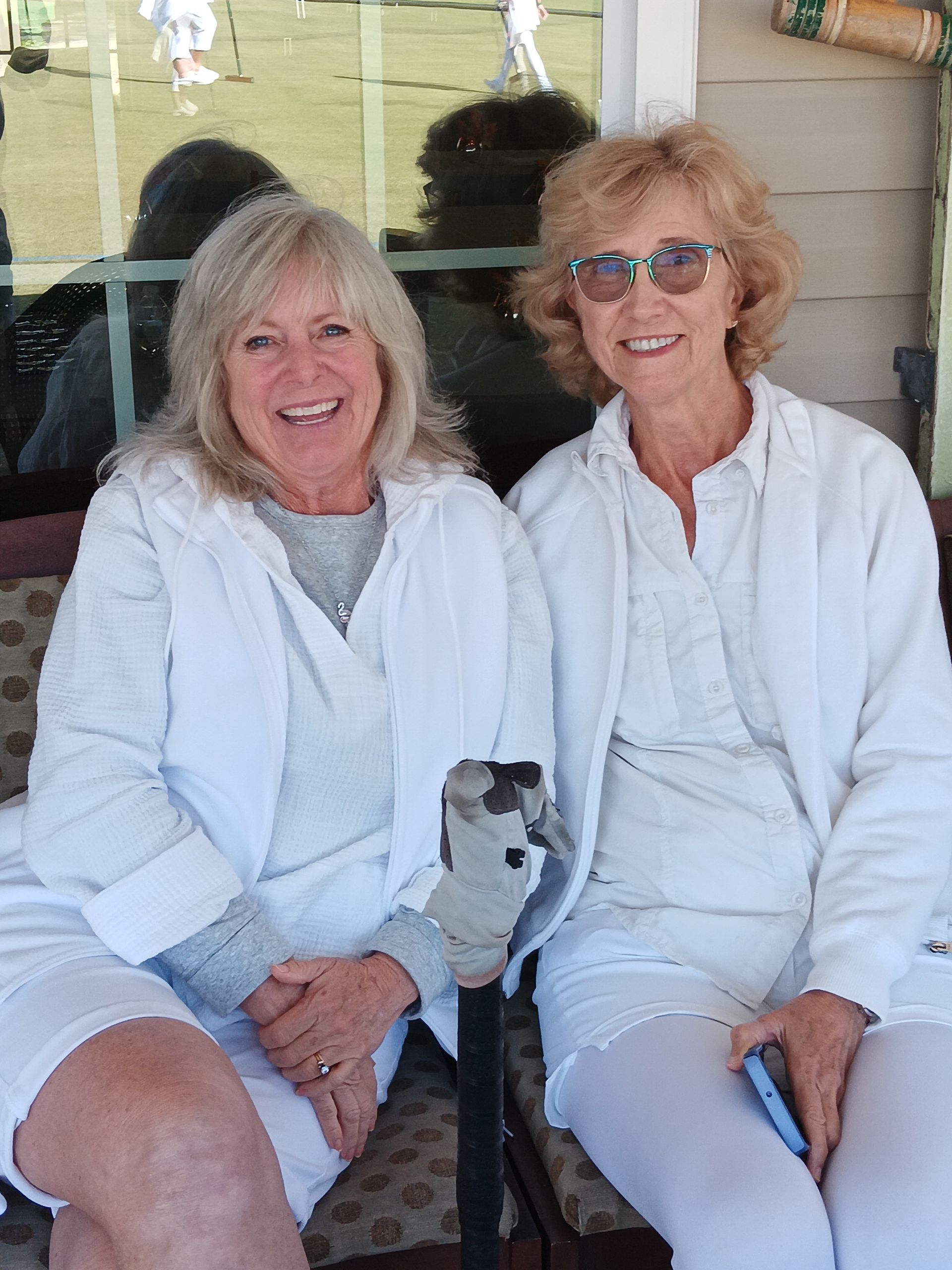 Two women in white outfits smile, sitting outdoors. One holds a cane.
