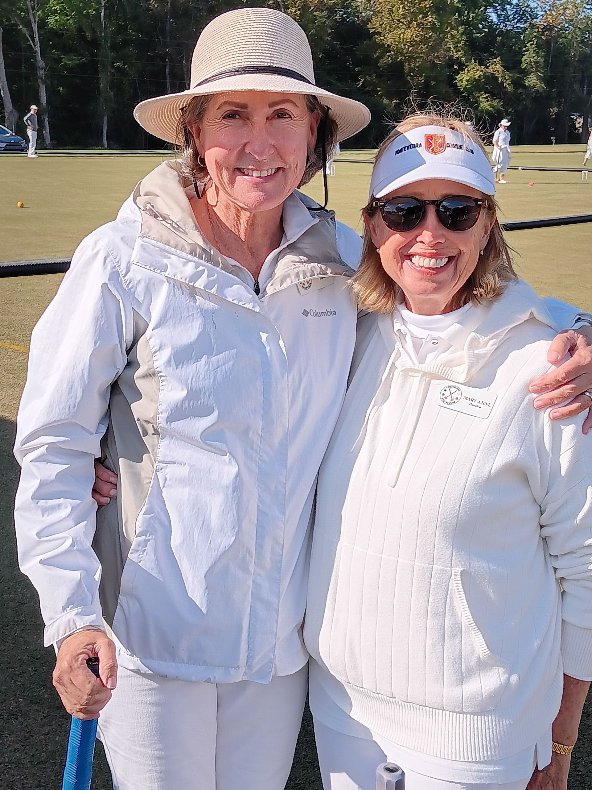 Two women smiling, arm-in-arm, on a croquet lawn. One wears a hat, the other sunglasses. Both are dressed in white.