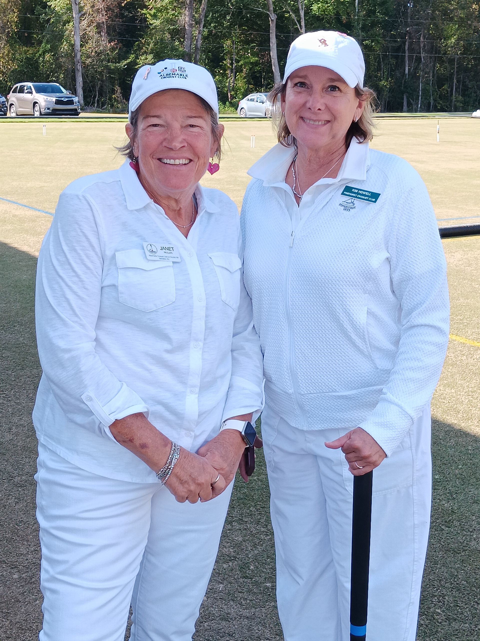Two women in white outfits and hats on a green field, one holding a croquet mallet.