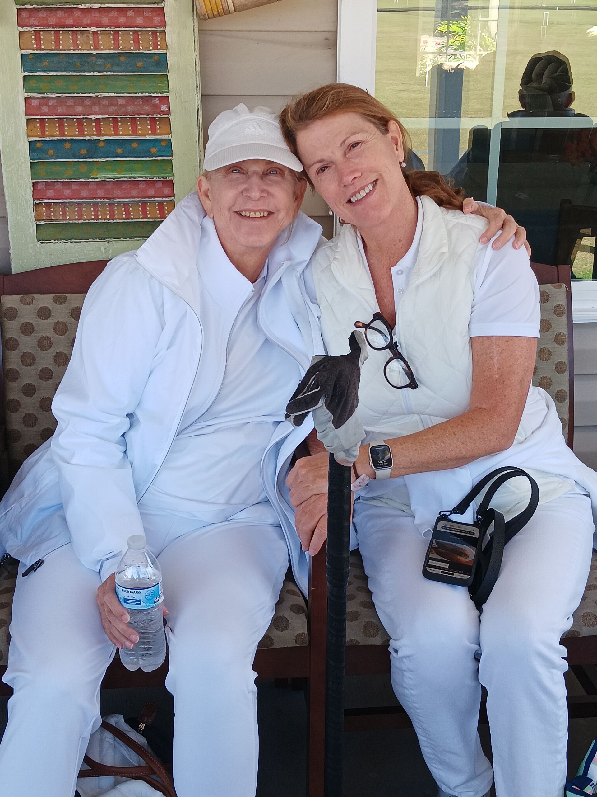 Two people smiling, sitting close together outdoors. One holds a cane, both wear white.