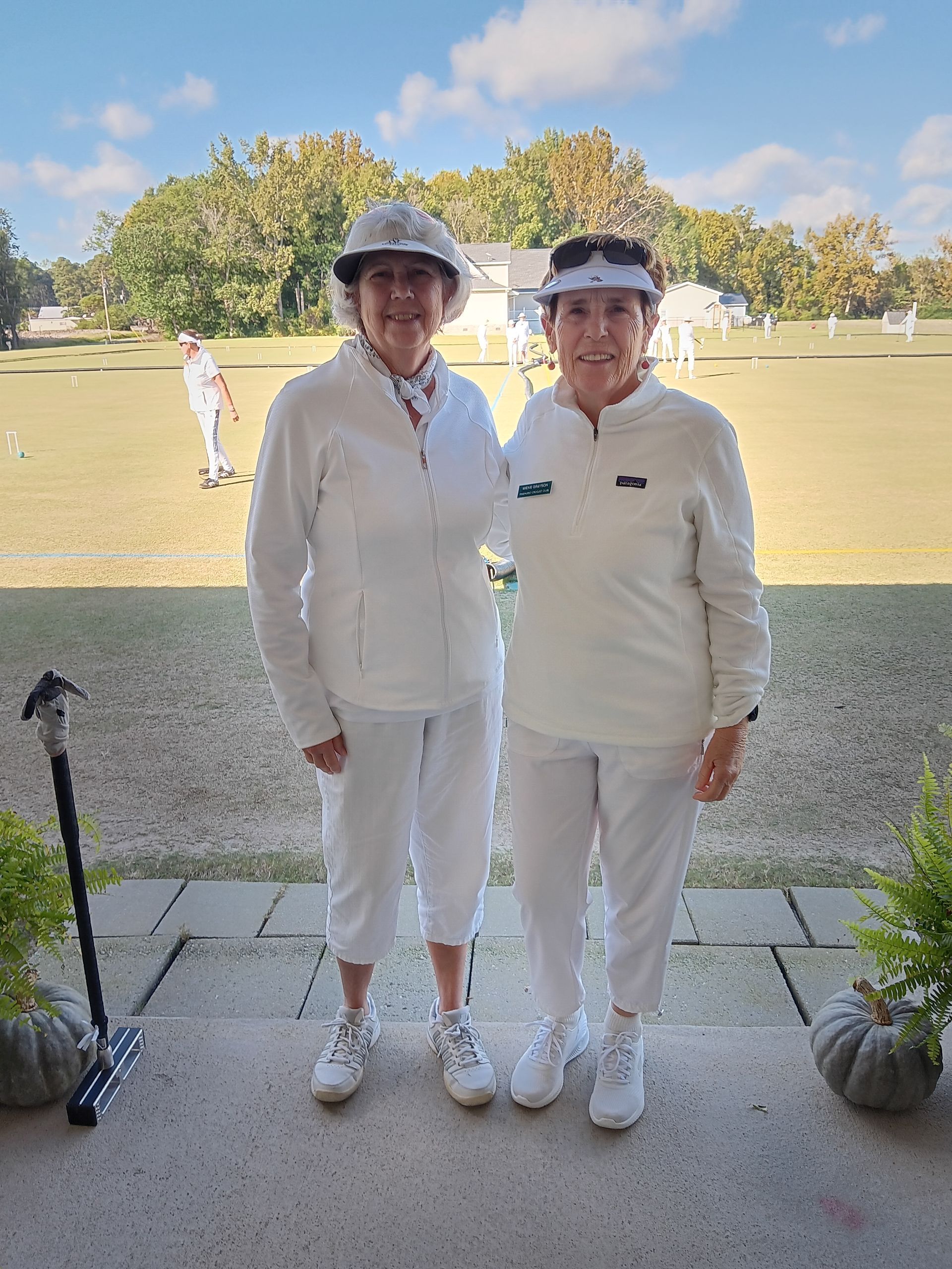 Two women in white outfits, standing on a patio; lawn bowling in background.