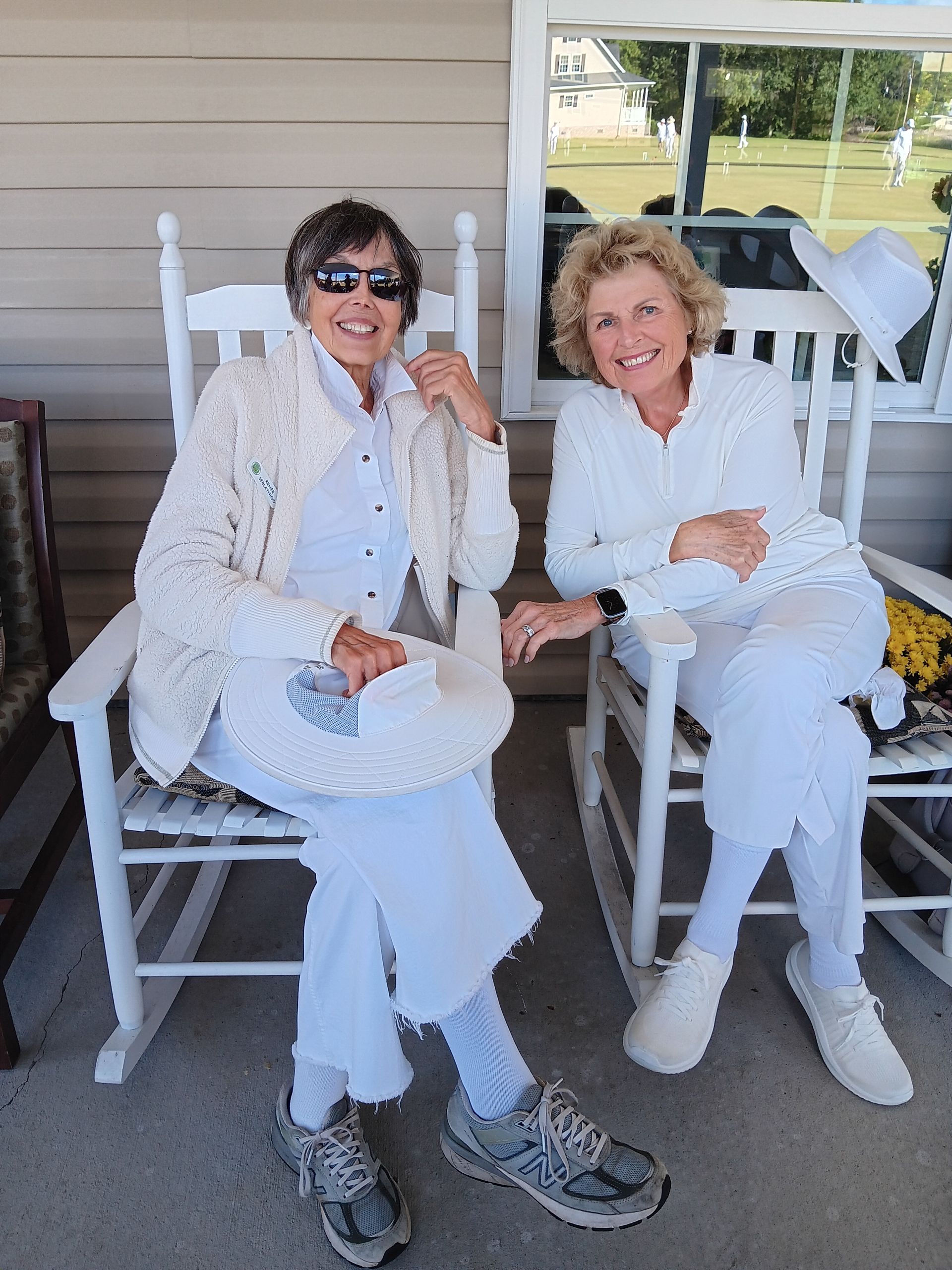Two women in white outfits smile while sitting in rocking chairs on a porch.