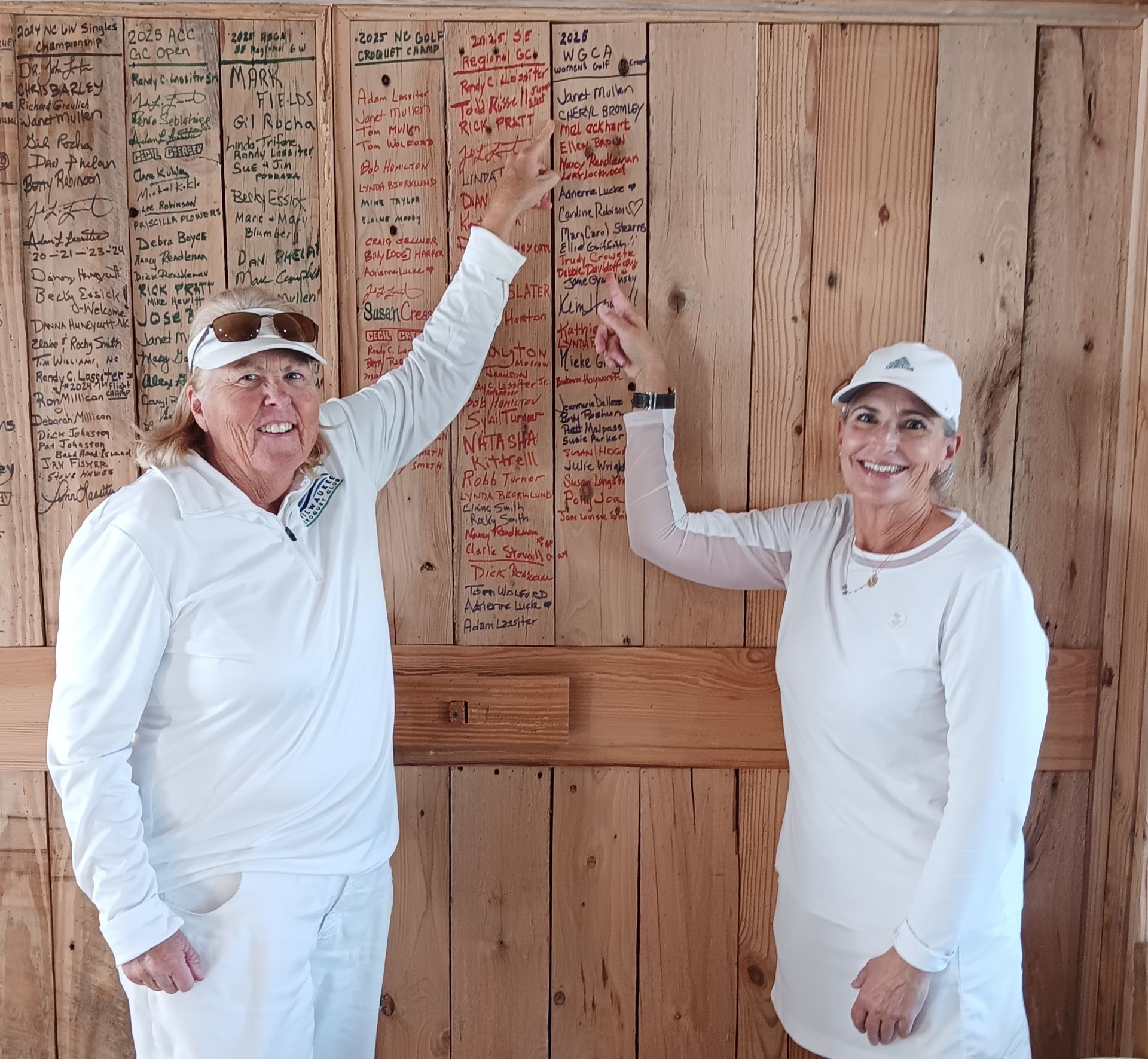 Two women in white tennis attire pointing at a wooden wall covered in handwritten notes.