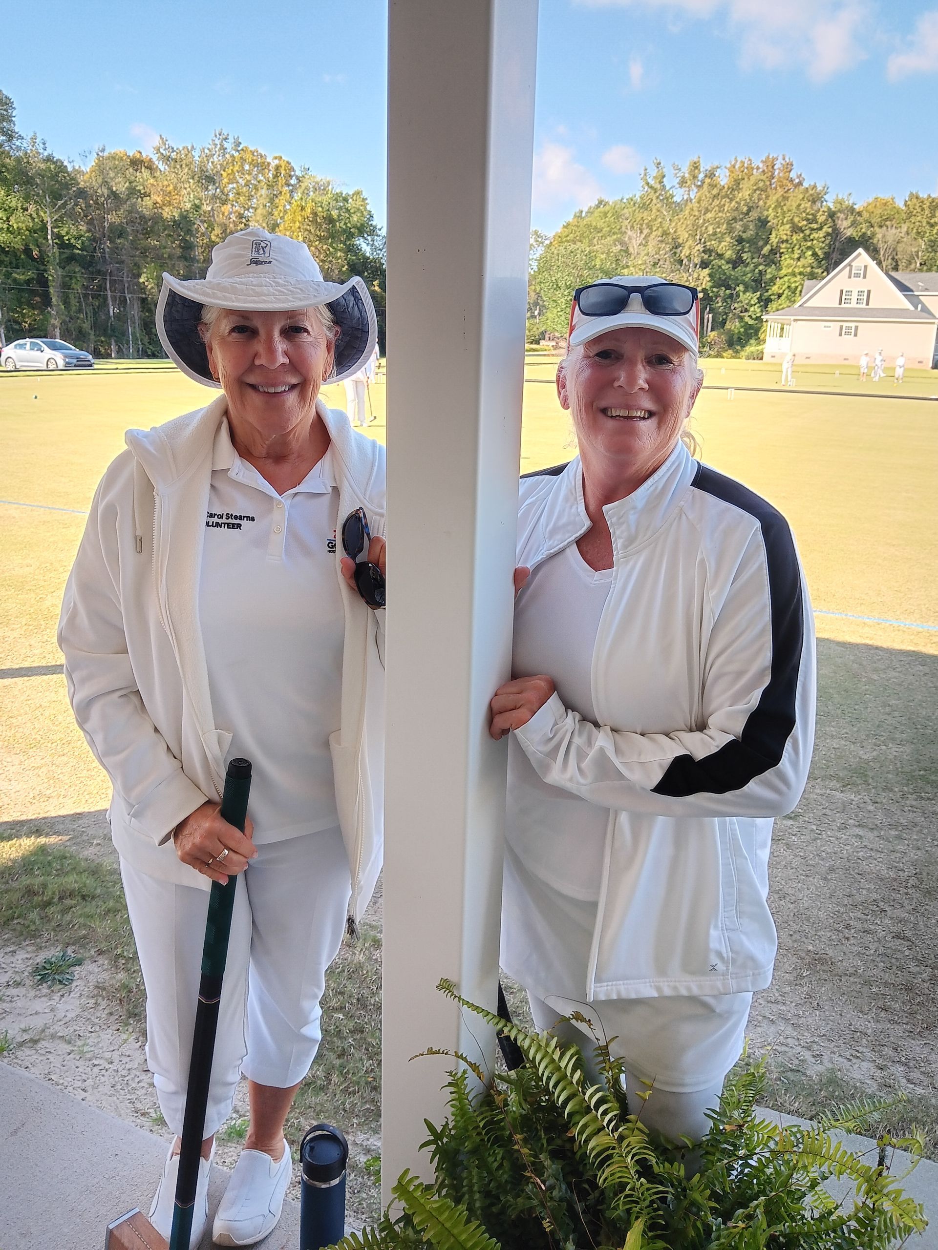 Two women in white outfits, one holding a croquet mallet, pose outdoors.