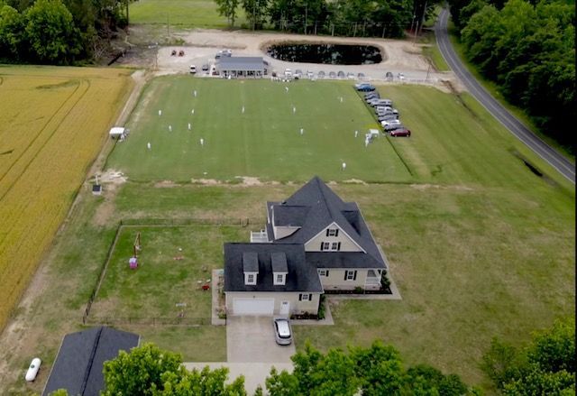 Aerial view of a house with a green lawn, sports field, and pond in a rural setting.