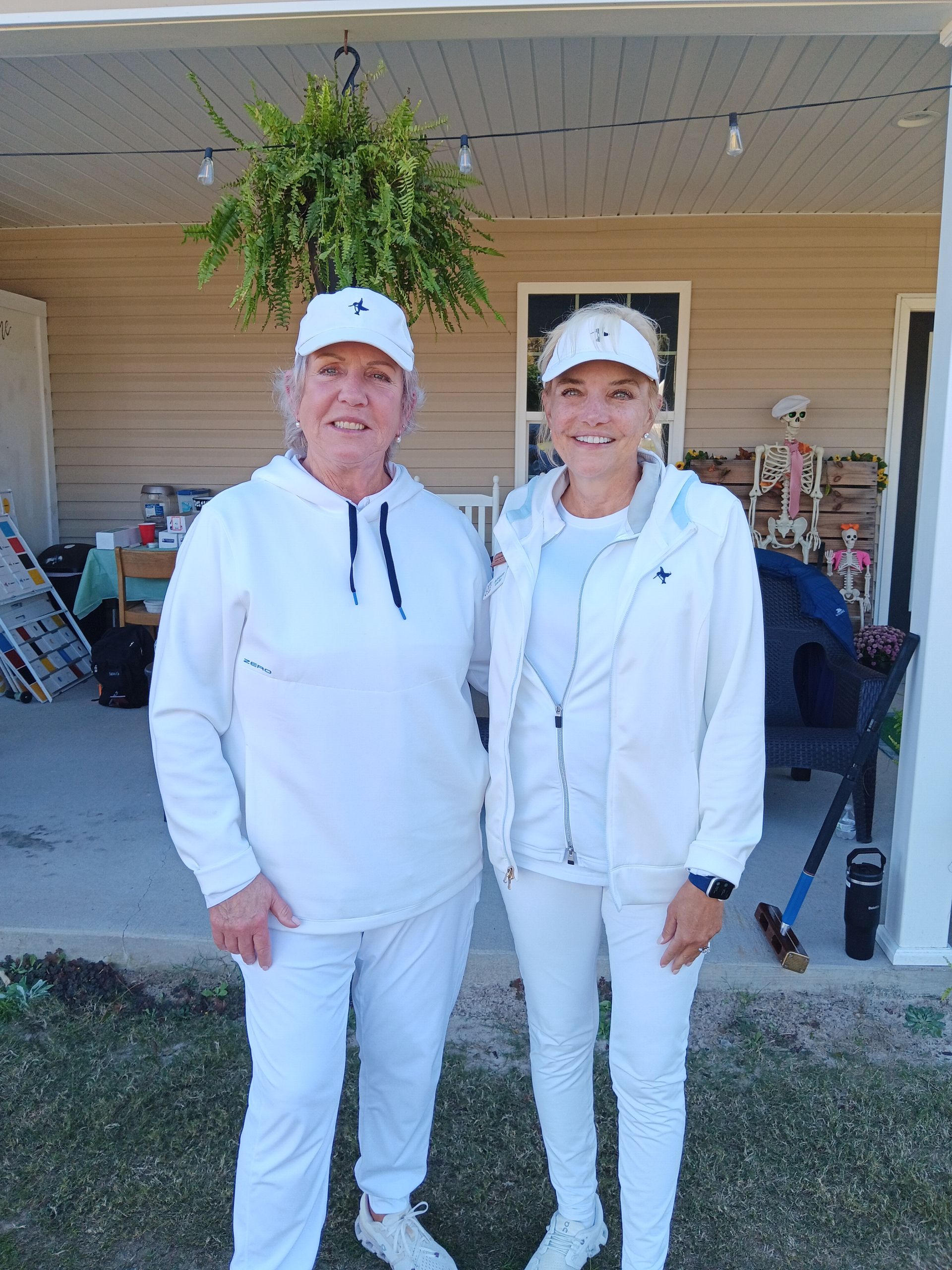 Two women in white outfits, standing on a porch. One wears a hoodie, the other a jacket. Fern hangs above them.
