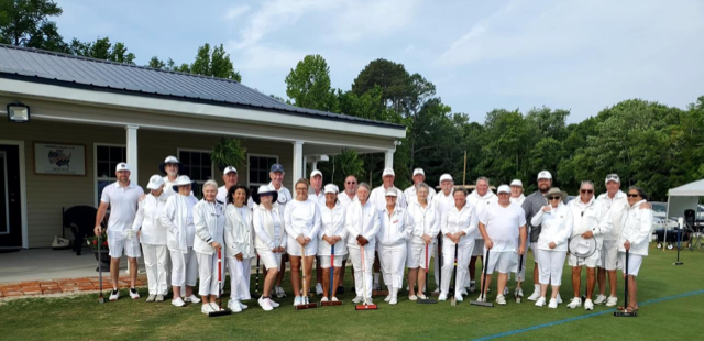 Group of people in white attire pose on a lawn, holding croquet mallets, in front of a building.